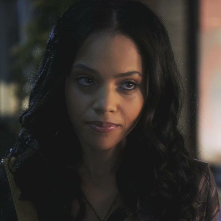 a woman with long dark hair and blue eyes looks at the camera while standing in front of a building