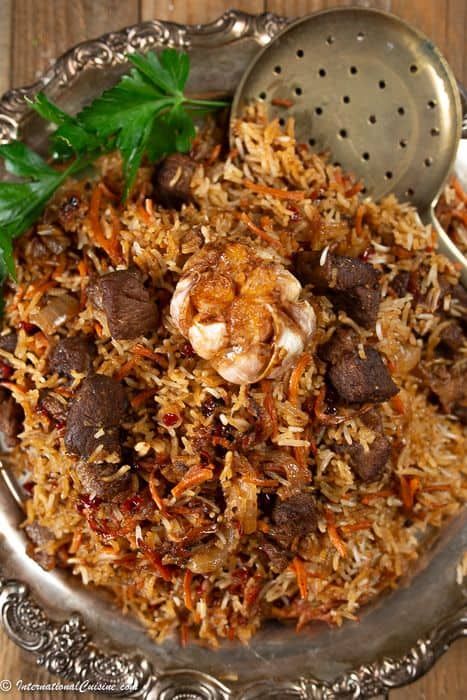 a silver bowl filled with rice and meat on top of a wooden table next to a metal strainer