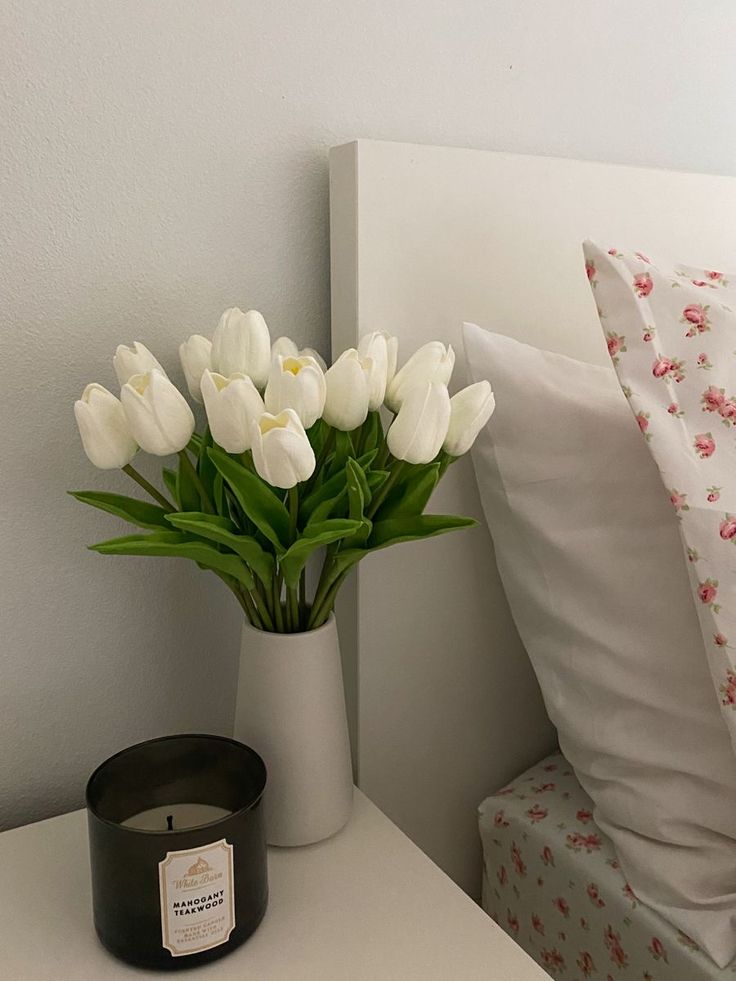 a vase with white tulips sits on a nightstand next to a candle