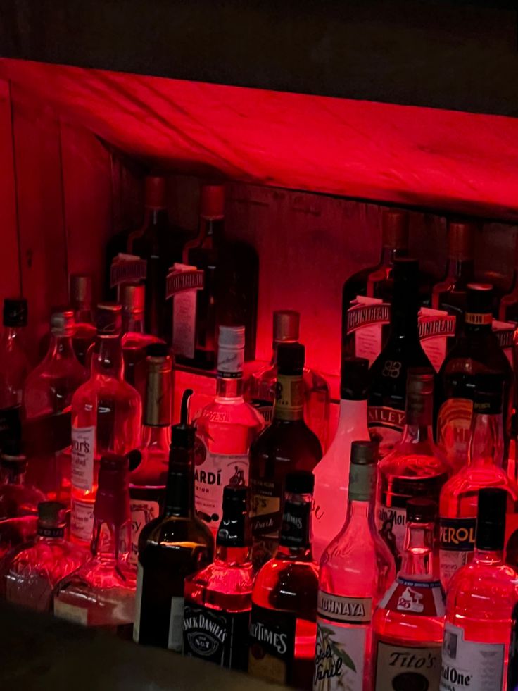 many bottles and glasses are lined up on the shelf in front of red light behind them