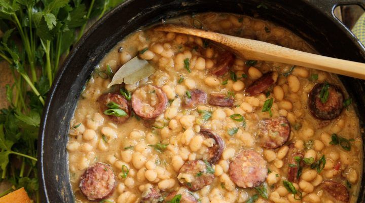 a pot filled with beans and sausages on top of a wooden spoon next to carrots