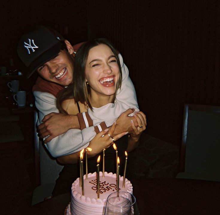 two people hugging each other in front of a pink cake with candles on it and one person wearing a baseball cap