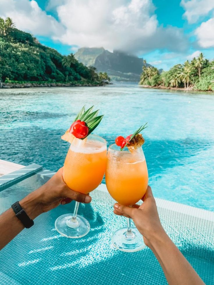 two people holding up glasses with drinks in front of water and palm trees on the beach