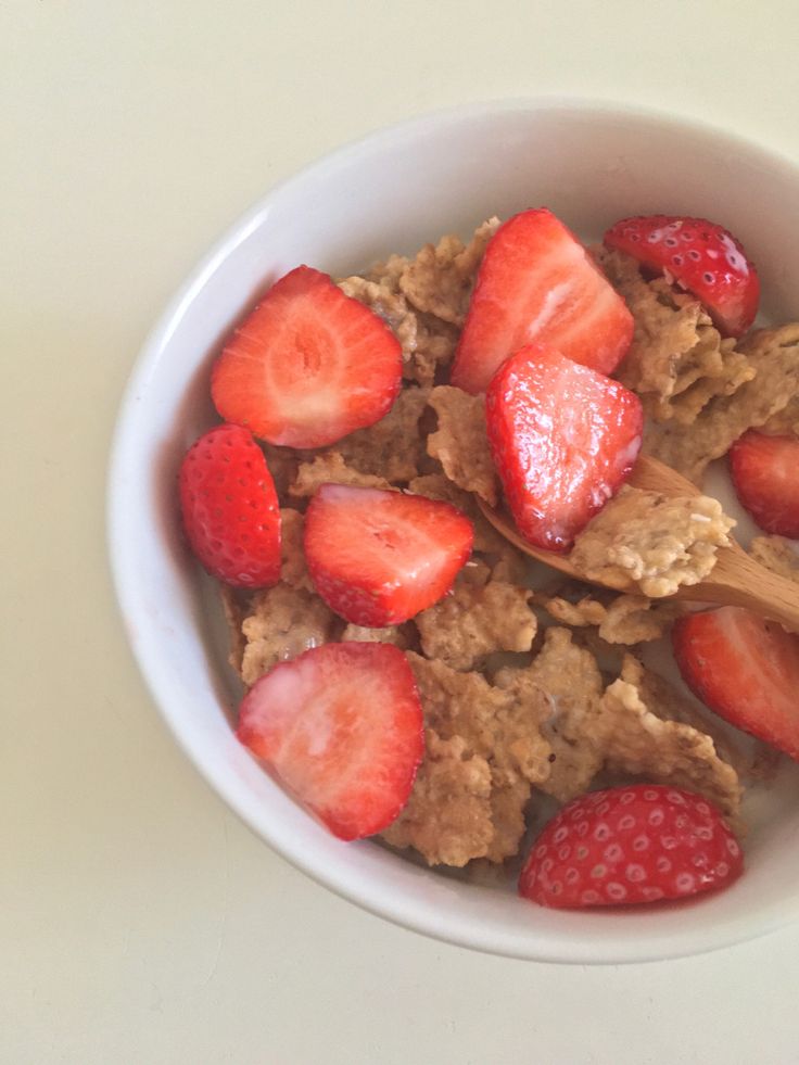 a bowl filled with cereal and strawberries on top of a white table next to a wooden spoon