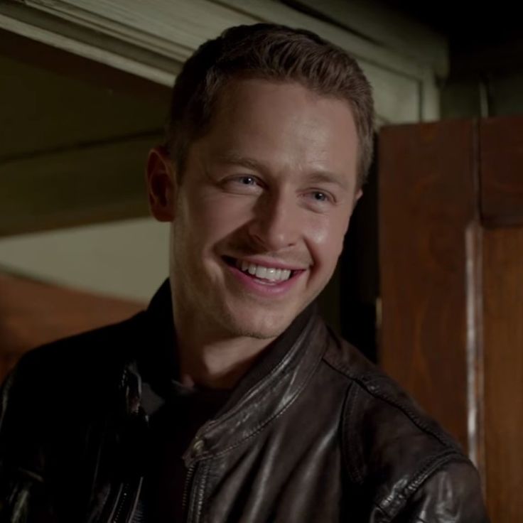 a smiling man wearing a leather jacket in front of a wooden paneled door and looking at the camera