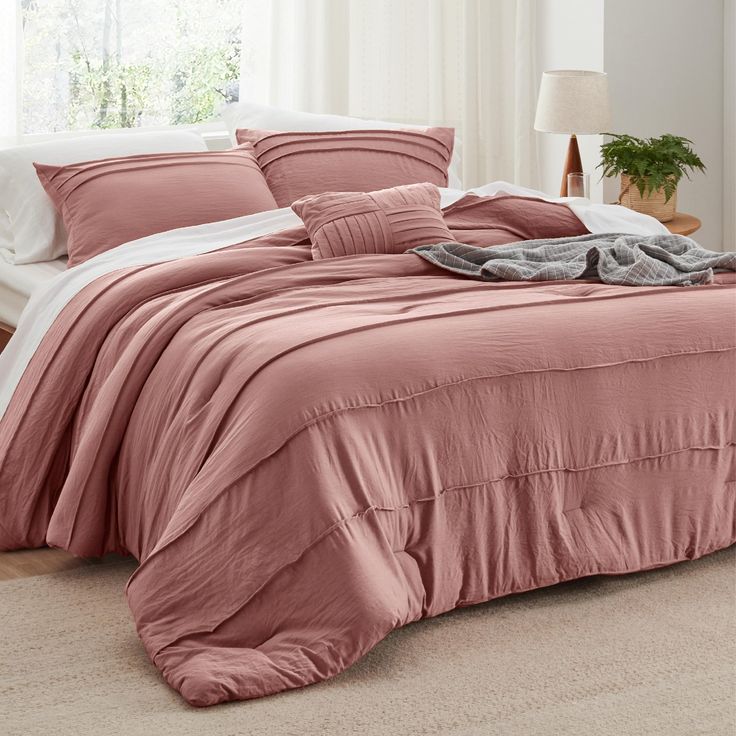 a bed with pink comforter and pillows in a white room next to a window