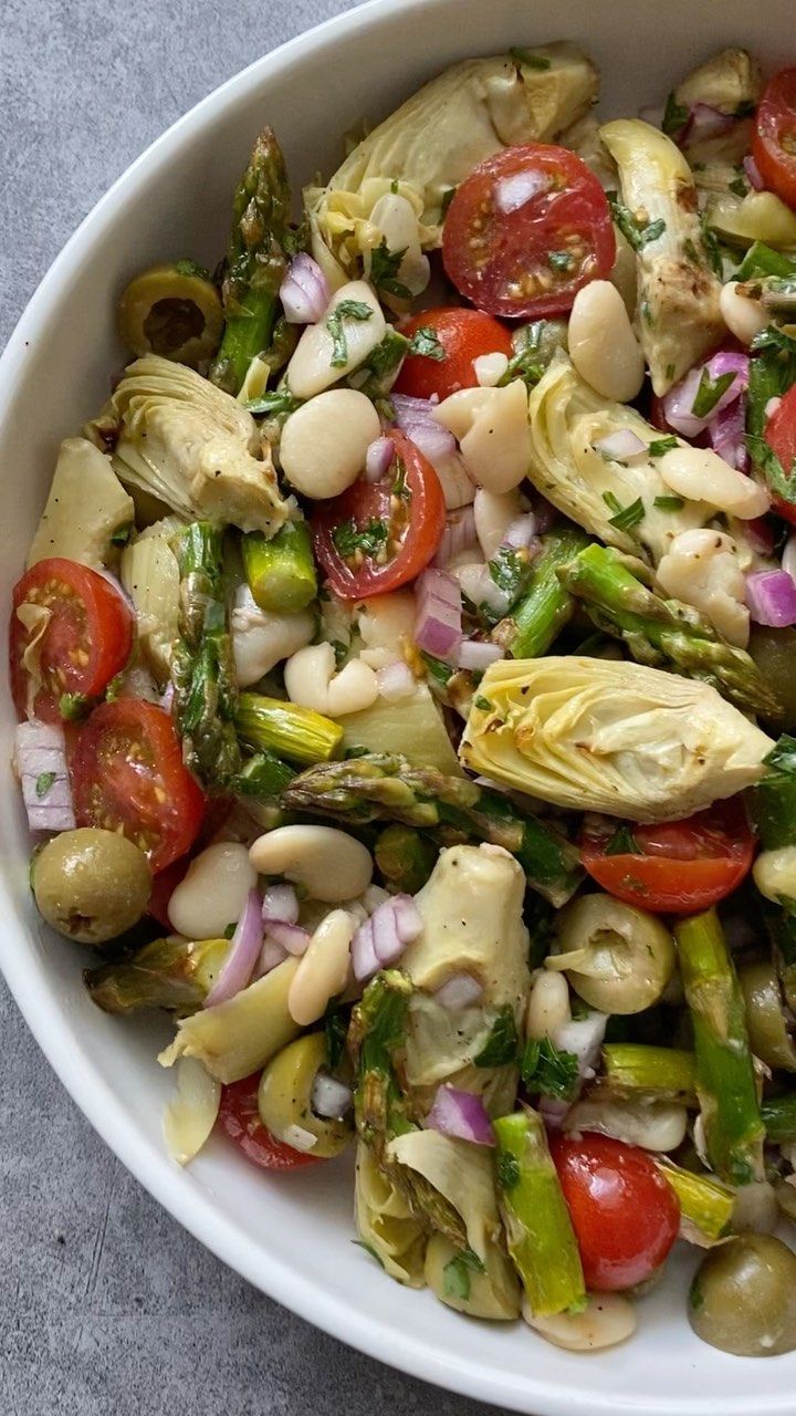 a white bowl filled with asparagus, tomatoes and artichokes on top of a table