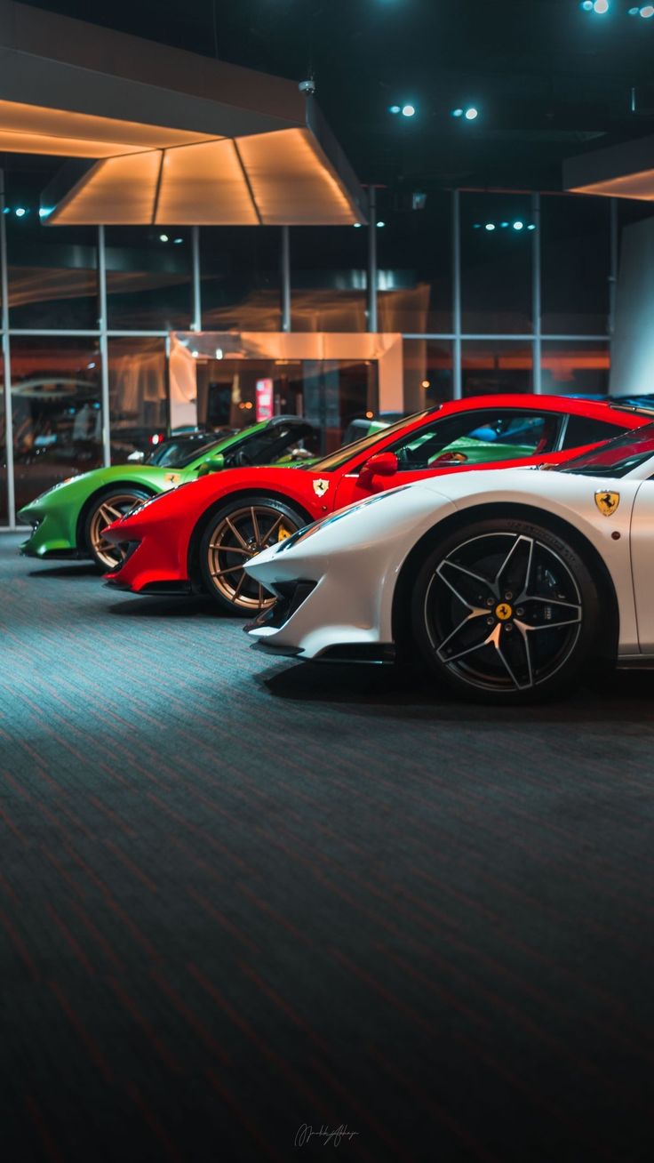 four different colored sports cars are parked in a showroom with large windows at night