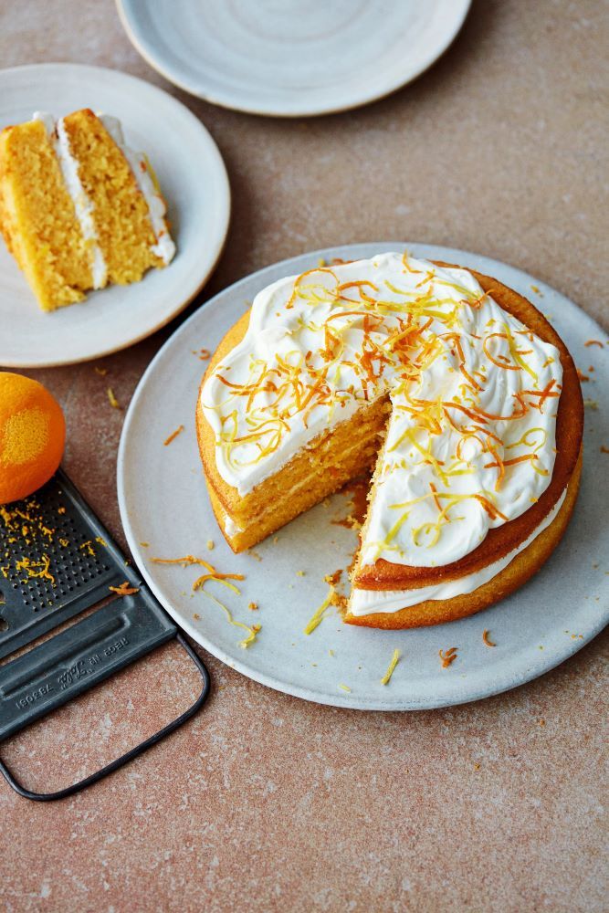 a cake with white frosting and orange zest is on a plate next to an orange slice