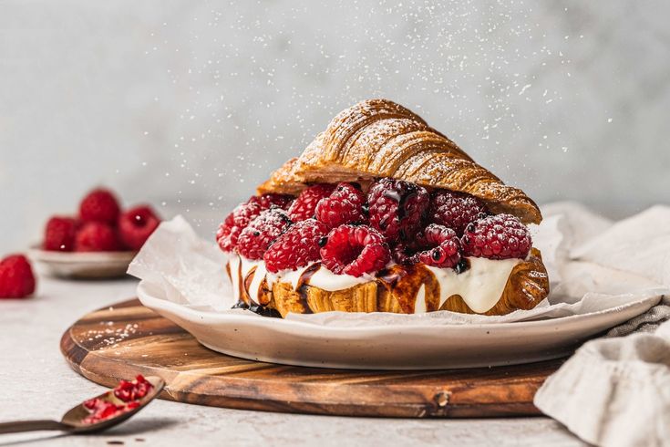 a croissant sandwich topped with raspberries and powdered sugar on a white plate