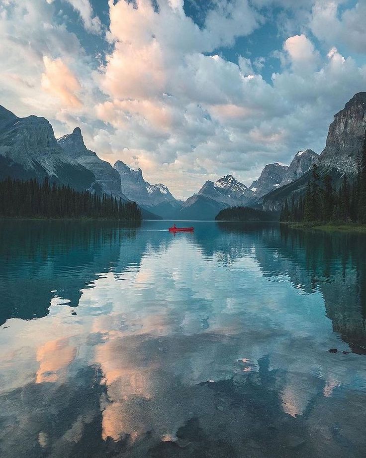 a lake surrounded by mountains and trees under a cloudy blue sky with red canoes in the water