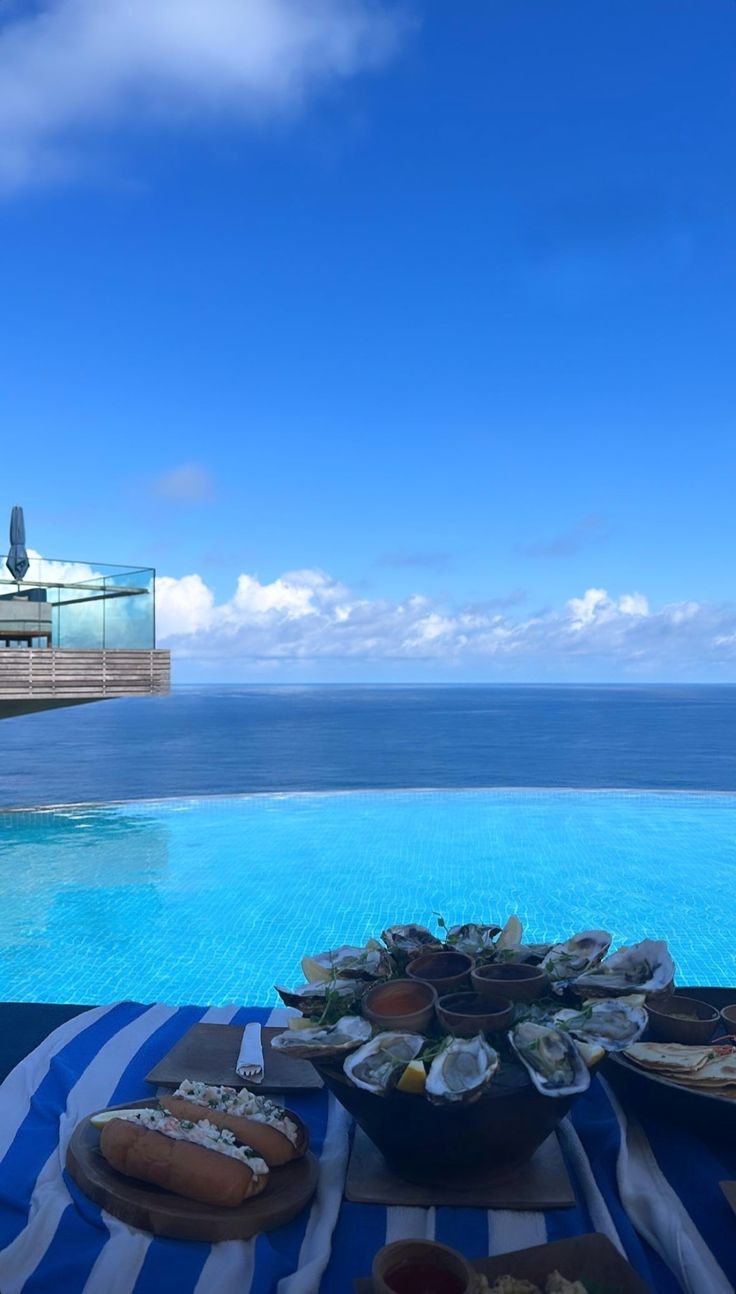 a table with plates of food on it next to a pool and the ocean in the background