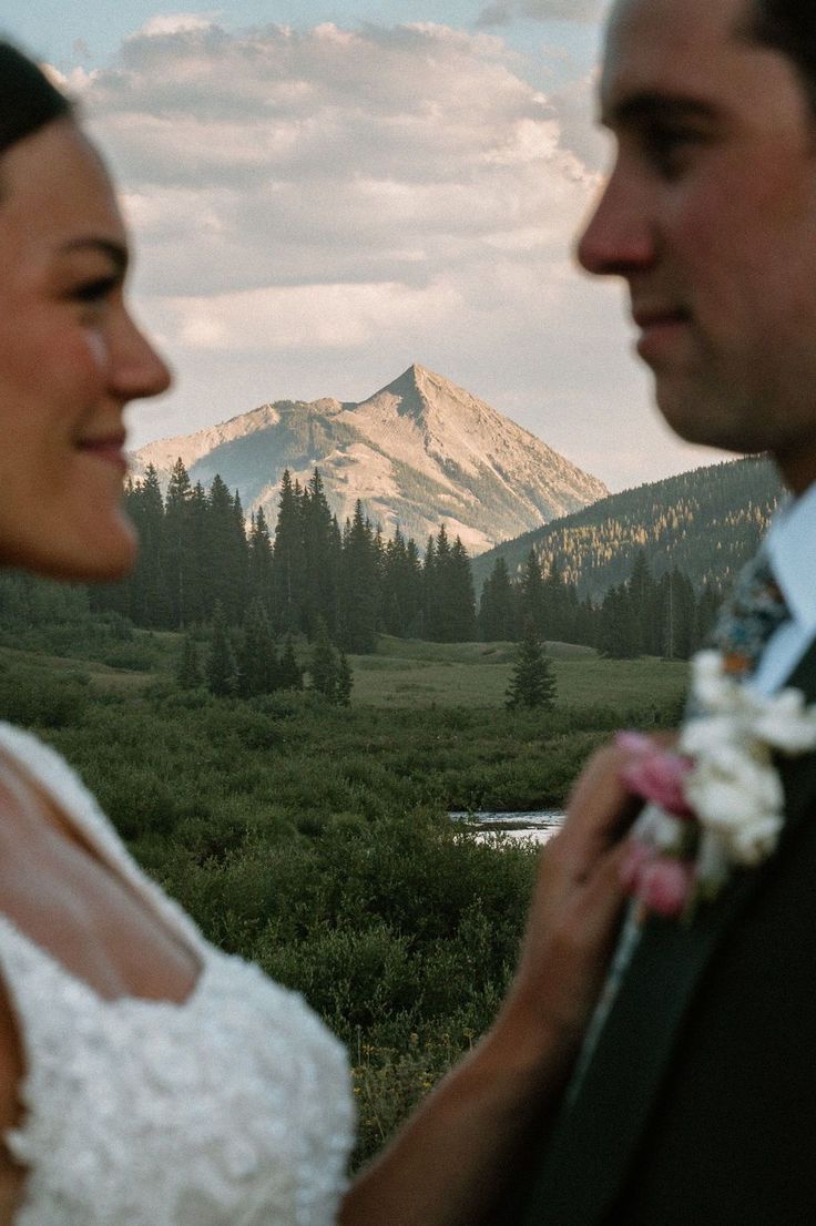 a bride and groom looking at each other in front of the mountains with flowers on their lap