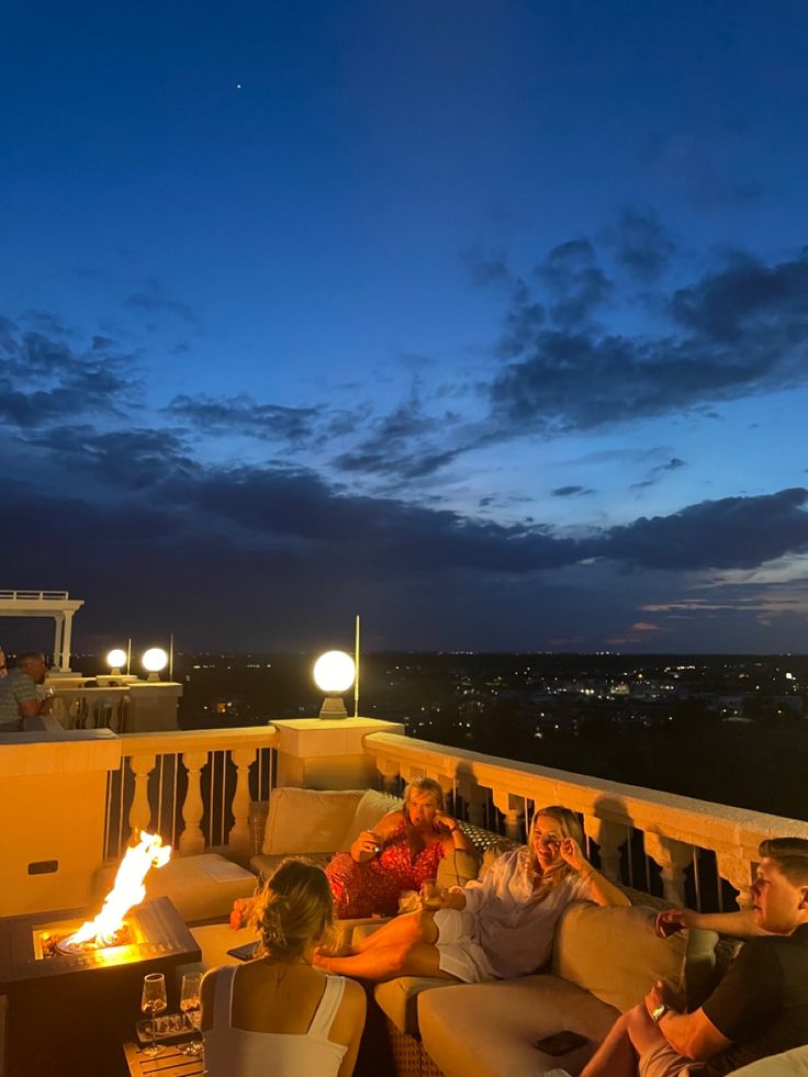 people are sitting on couches around a fire pit at night with the city lights in the background