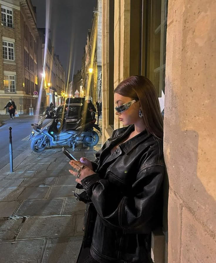 a woman leaning against a wall looking at her cell phone while wearing sunglasses and leather jacket