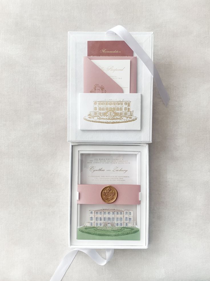 two wedding cards in a white box with pink ribbon and a gold seal on top