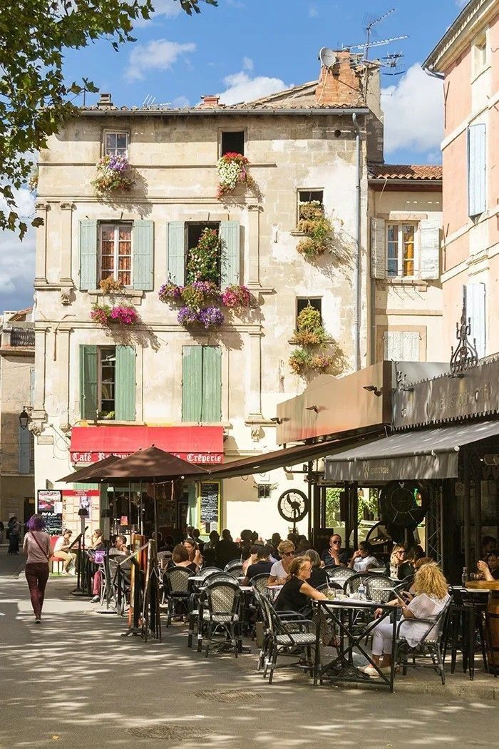 people are sitting at tables in an outdoor cafe on a sunny day with flowers hanging from the windows