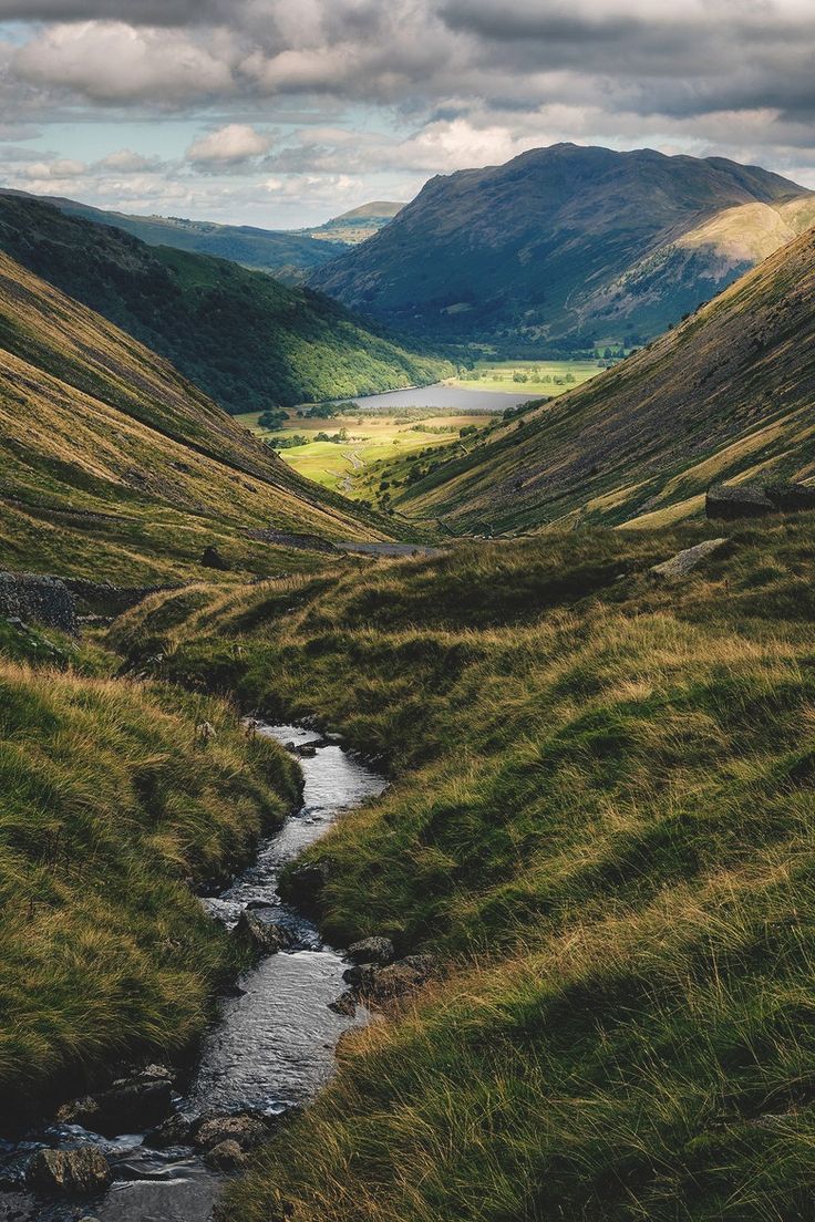 a small stream in the middle of a grassy valley with mountains in the background and cloudy skies above