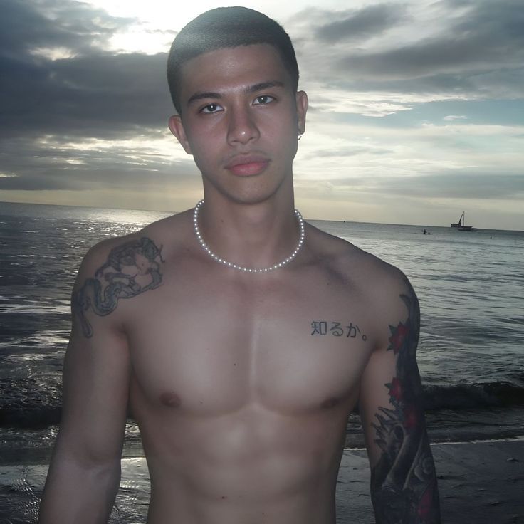 a shirtless man standing in front of the ocean with tattoos on his arms and chest