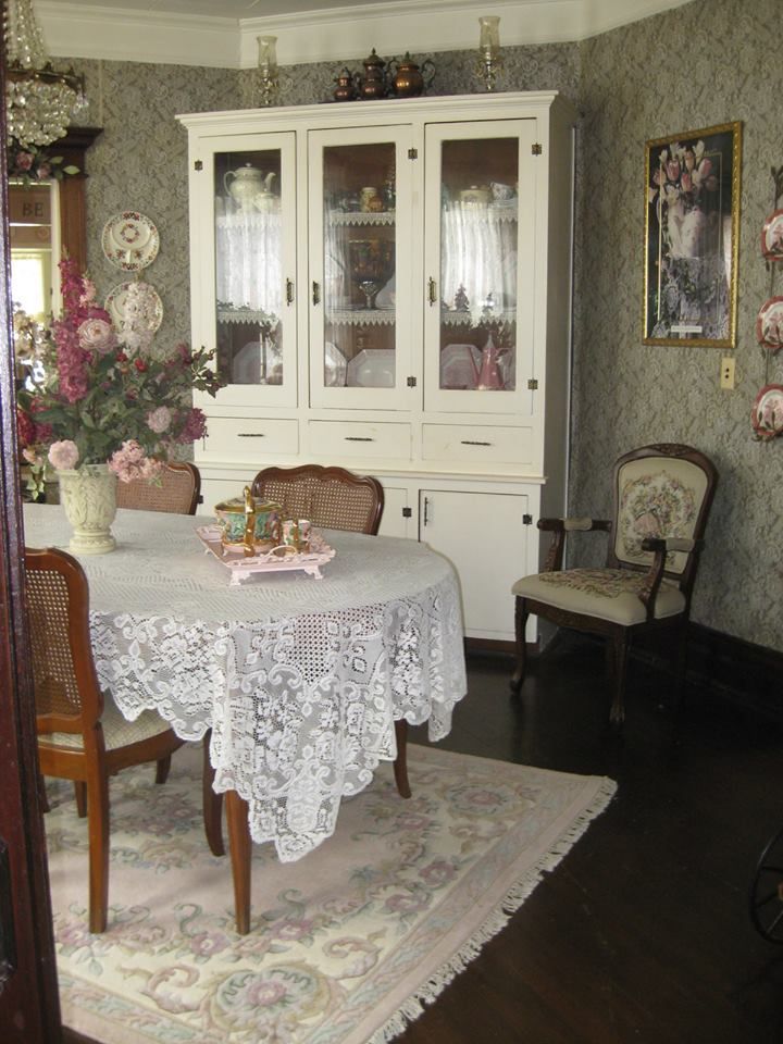 a dining room table and chairs with white lace on the table cloth, in front of a china cabinet