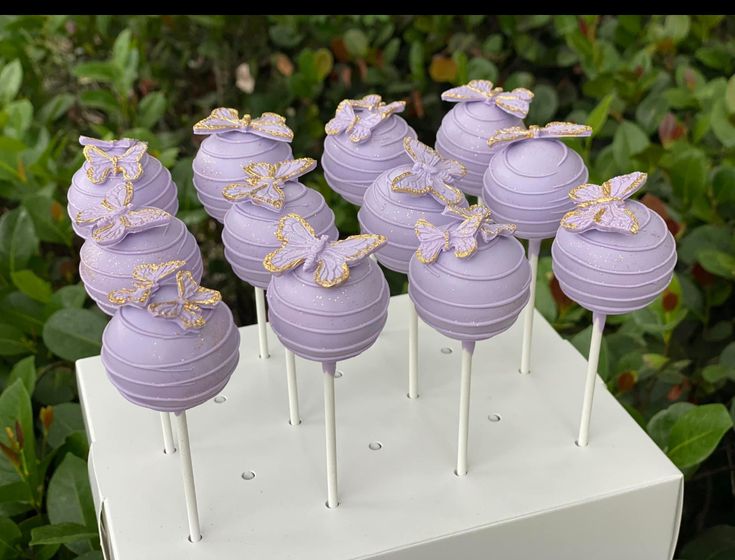 there are many purple cake pops with bows on them sitting on a white stand in front of some bushes