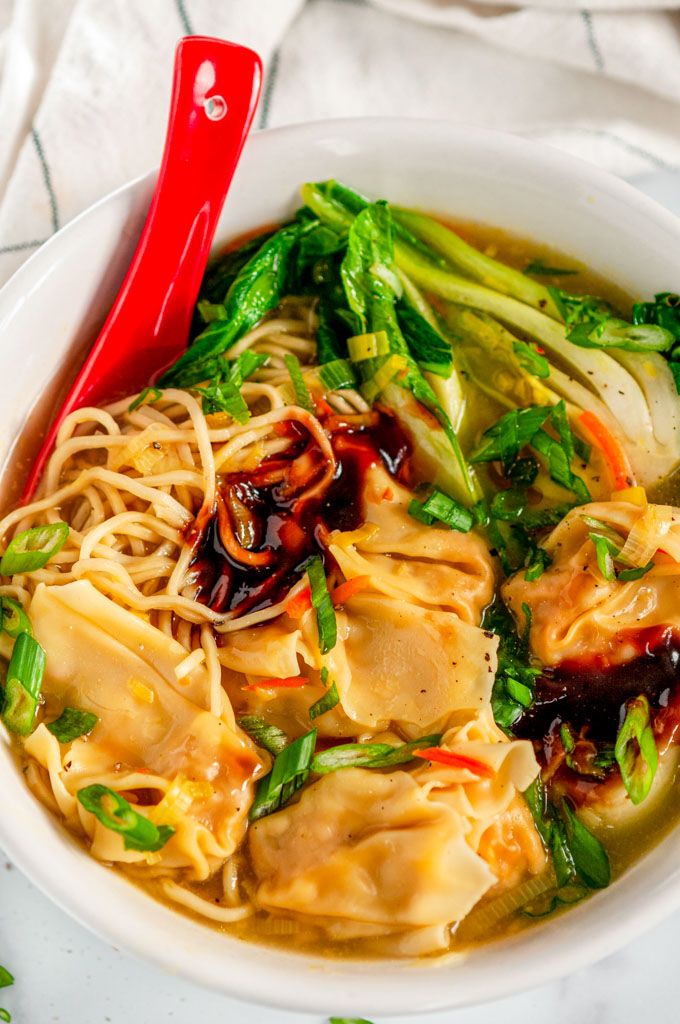 a white bowl filled with noodles and veggies next to a red spoon on top of a table