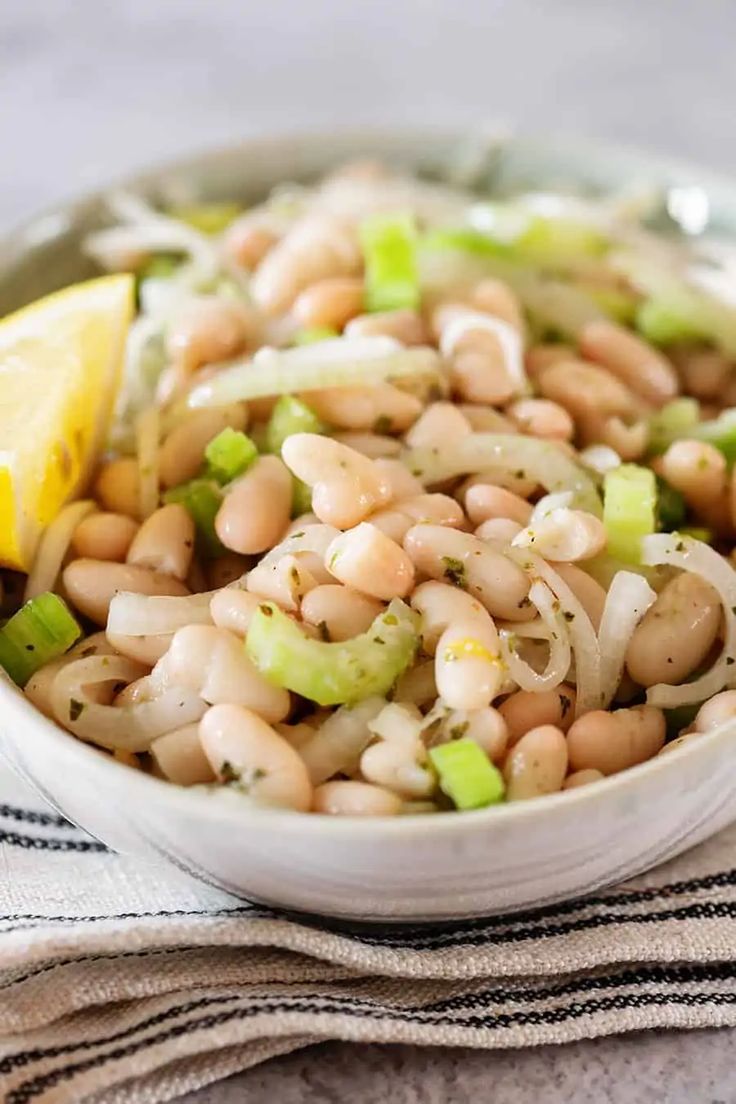 a white bowl filled with beans, onions and celery next to a lemon wedge