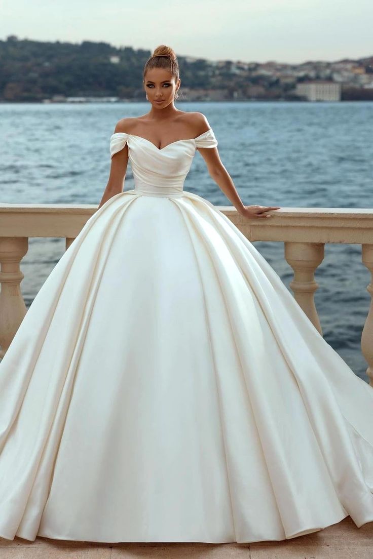 a woman in a white wedding dress standing on a balcony near the water with her hands on her hips