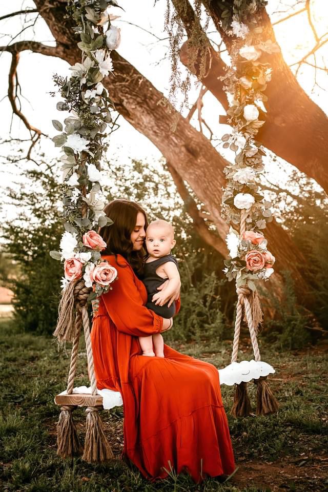 a woman holding a baby in her arms while sitting on a swing with flowers hanging from it