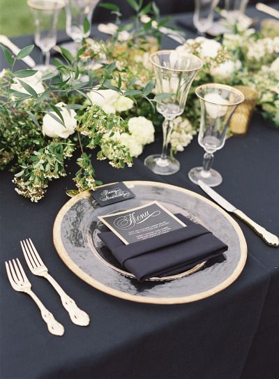 a black table cloth with white flowers and greenery on it is set for a formal dinner