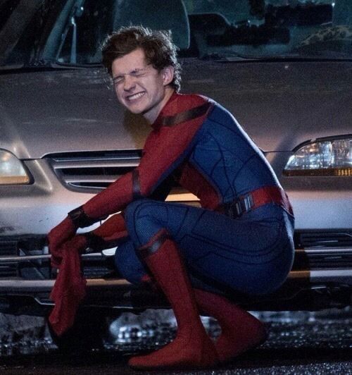 a man dressed as spider - man sitting on the ground next to a silver car