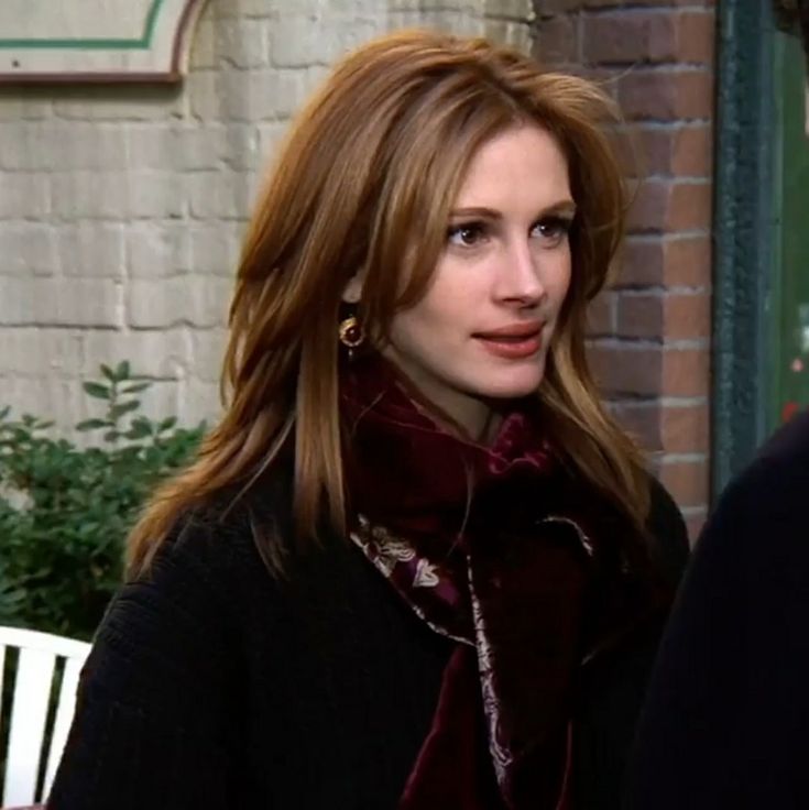 a woman standing next to a brick wall wearing a black coat and maroon scarf with gold earrings