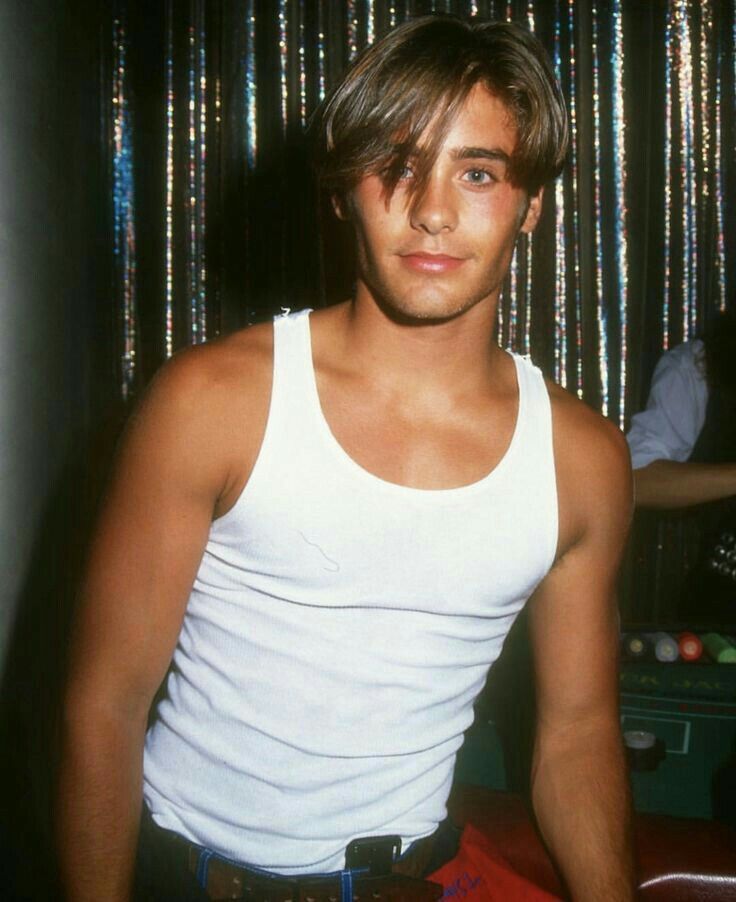 a young man wearing a white tank top standing in front of a wall with tinsel on it