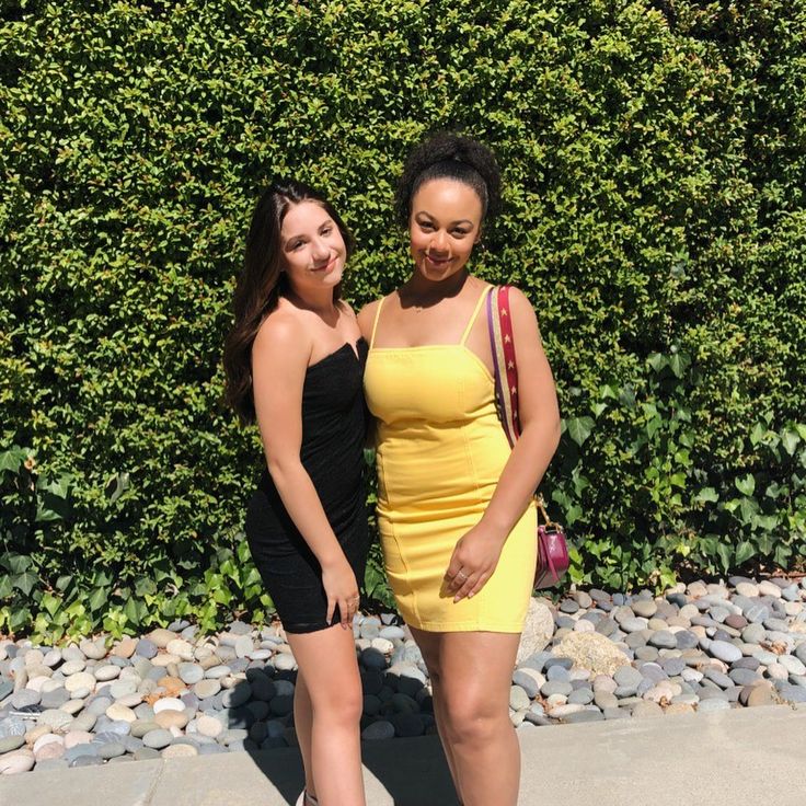 two young women standing next to each other in front of a green wall and bushes