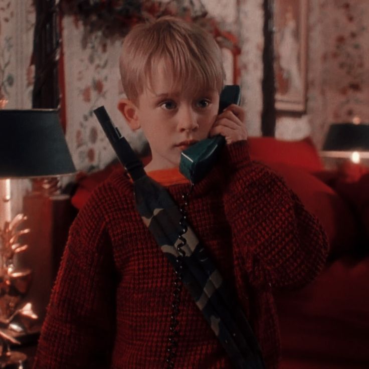 a young boy holding a phone to his ear while standing in front of a christmas tree