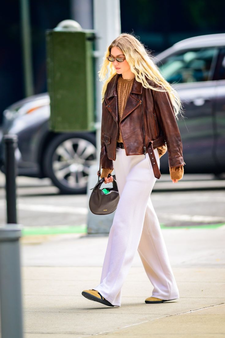 a woman walking down the street in white pants and a brown jacket with her hair blowing back