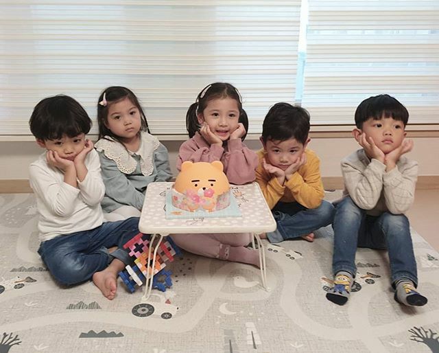 five children sitting on a bed with a cake in front of them and one child holding a teddy bear