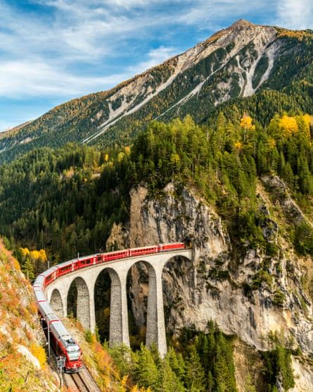 a red train traveling over a bridge in the middle of a mountain range with fall foliage