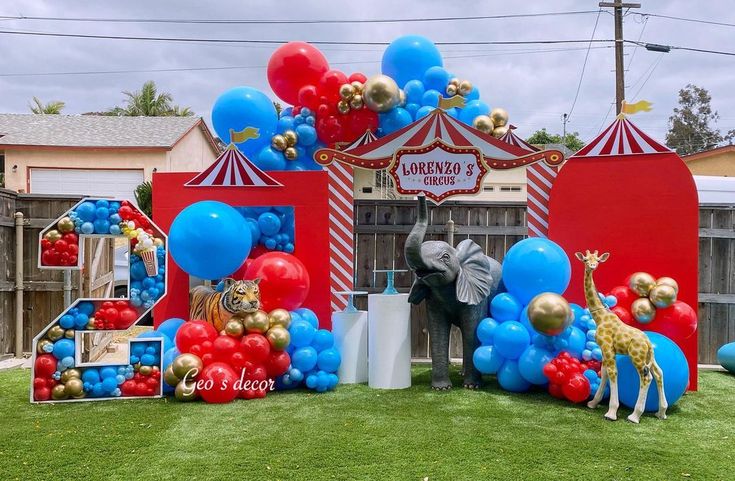 an elephant and giraffe stand in front of a circus tent with balloons on the grass