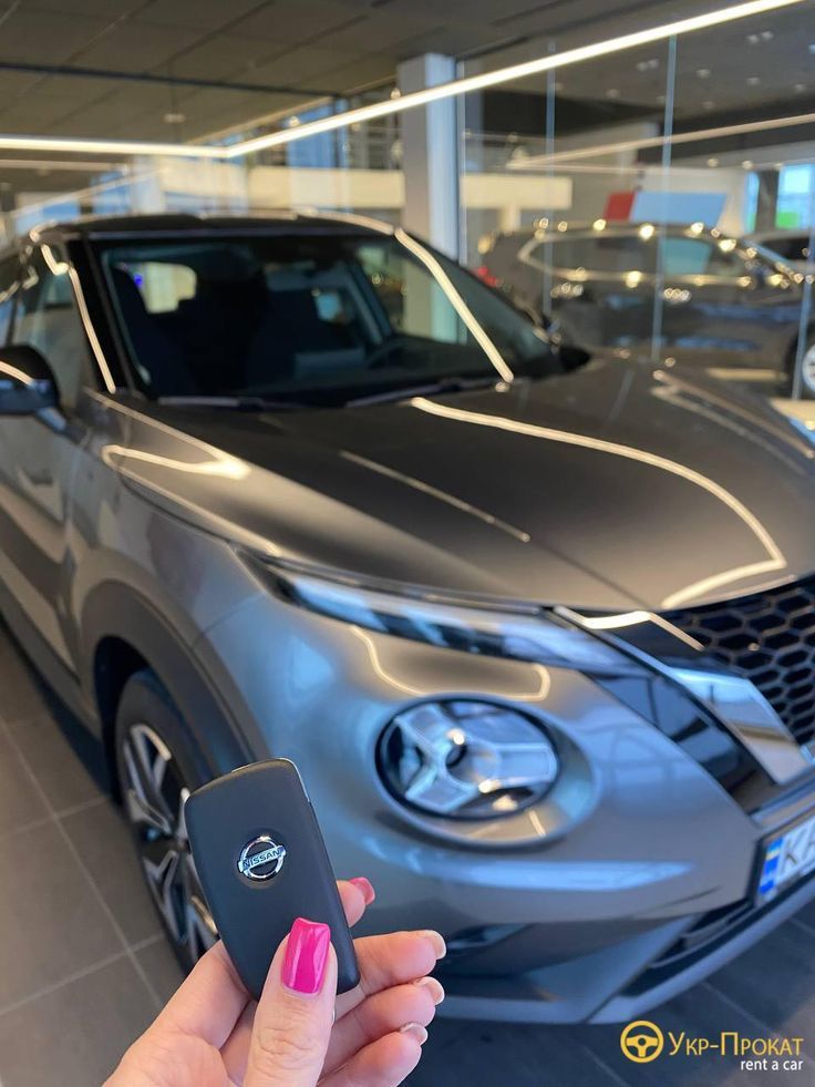 a woman holding a cell phone in front of a gray nissan car at a dealership