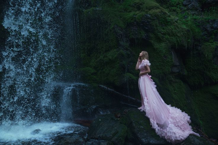 a woman in a pink dress standing next to a waterfall