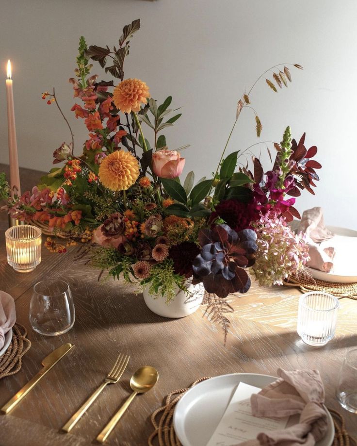 an arrangement of flowers in a vase on a table with candles and napkins around it