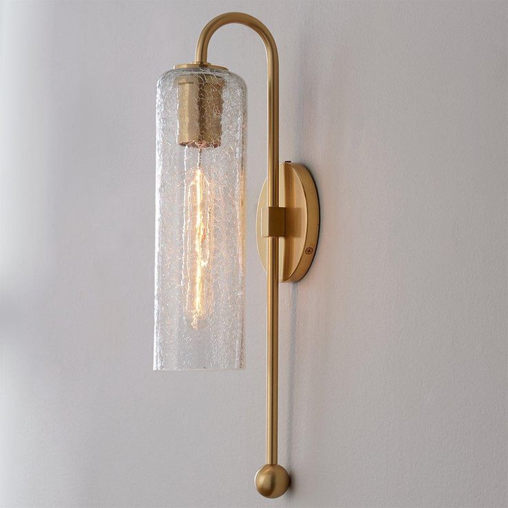 a wall light with a clear glass shade on the top and gold metal arm, against a white wall