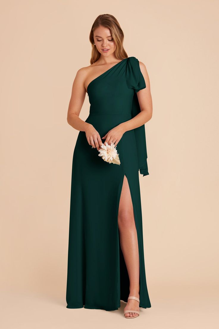 a woman in a dark green dress with one shoulder and slited skirt, holding a flower