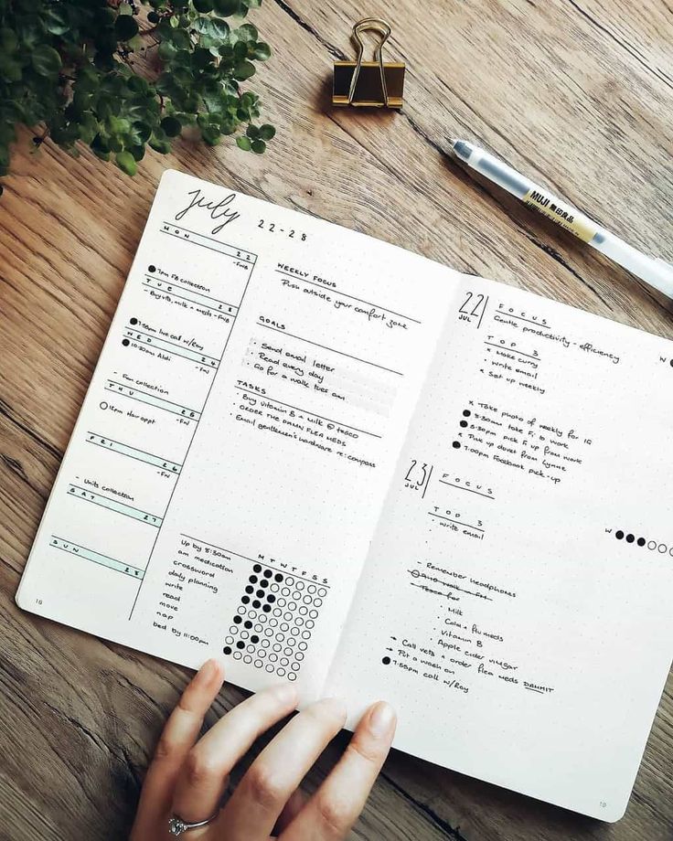 a person's hand is on top of an open notebook next to a pen and plant