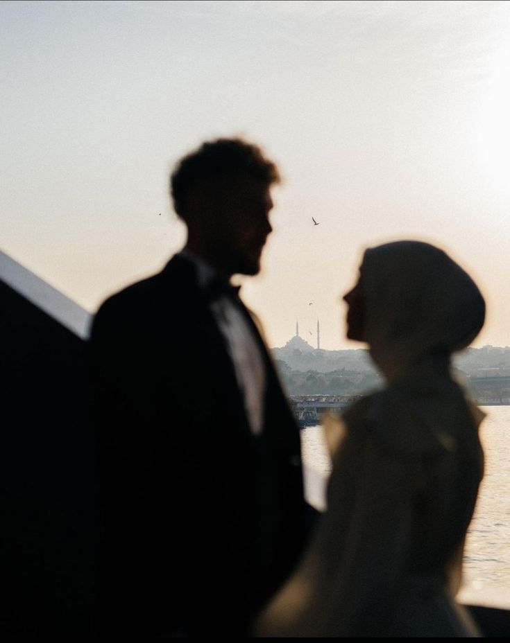 two people standing next to each other in front of a body of water at sunset