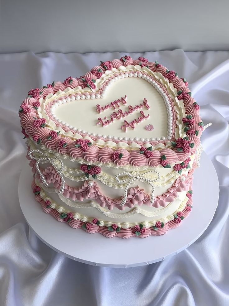 a heart shaped cake with pink and white frosting sitting on a tablecloth covered surface