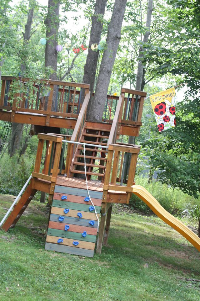 a wooden play set in the middle of a wooded area with a slide and climbing wall