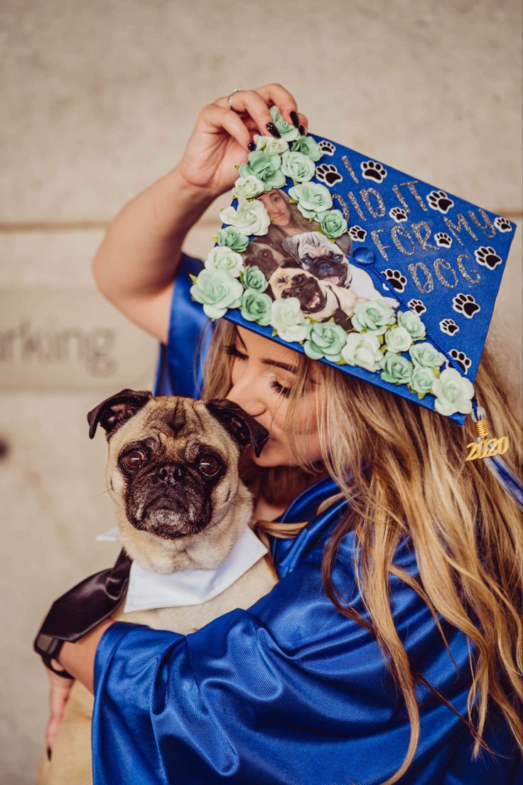 a woman in a blue graduation gown holding a pug wearing a hat with flowers on it