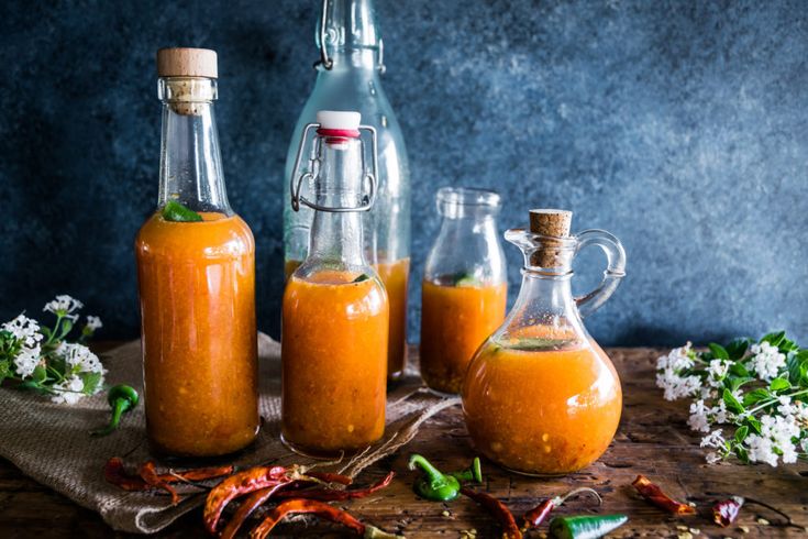 three glass bottles filled with orange liquid next to some chili peppers and flowers on a wooden table